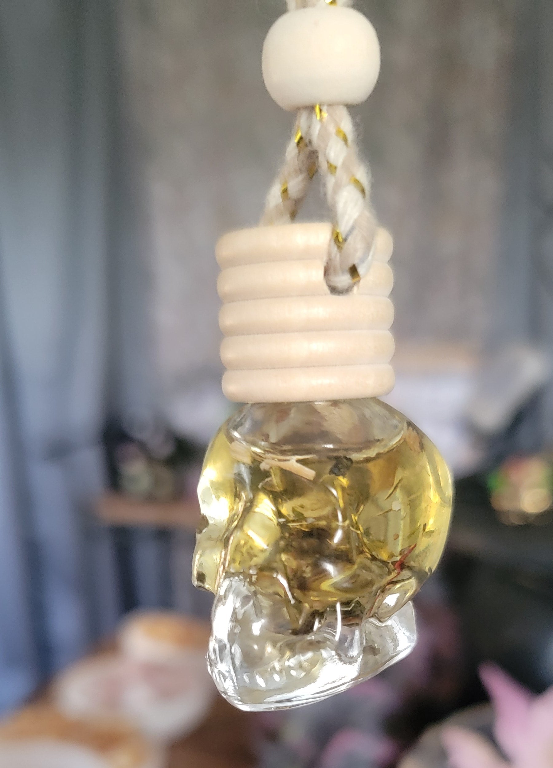 A glass skull-shaped car air freshener with a yellow green tint, hanging from a metal chain, with crystal and herbs inside the skull and a cork lid attached to the top.