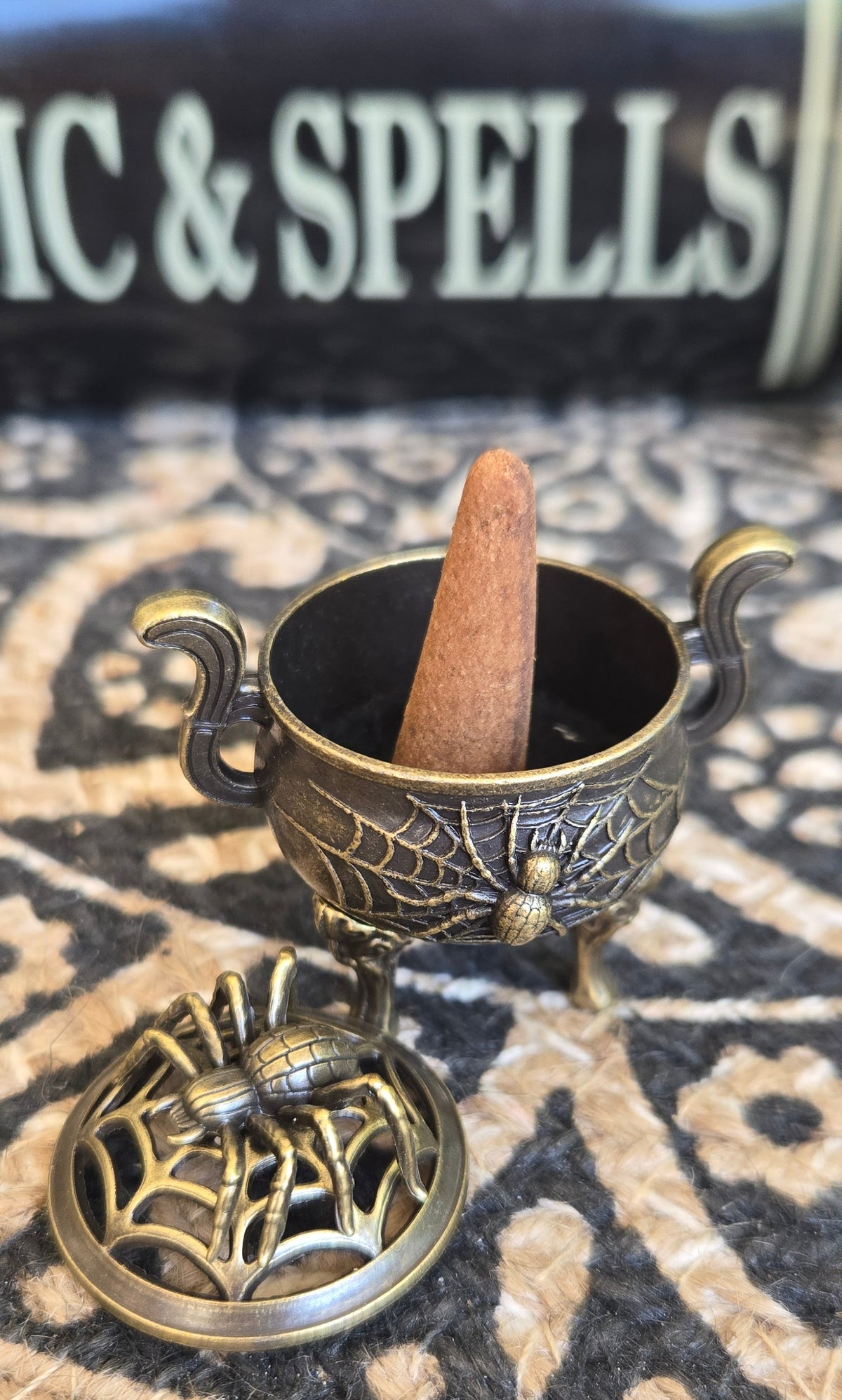 A small ornate brass incense burner with a matching lid, featuring spiderweb detailing and a conical incense stick inside, placed on a patterned rug in front of a book titled 'Magic & Spells'.