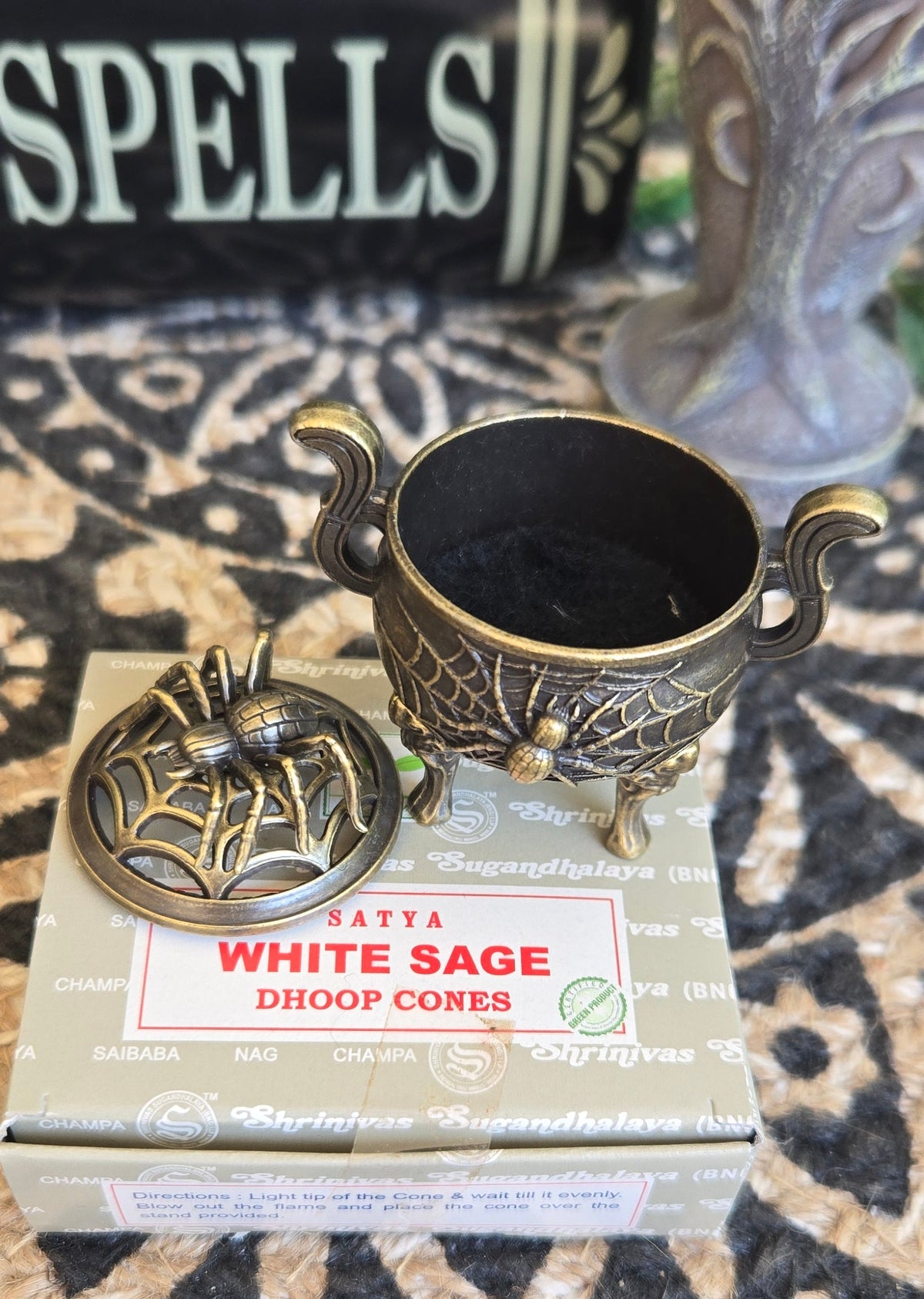 A brass-colored metal diyoop cone holder with a lid, placed on top of its product box labeled 'SATTVA WHITE SAGE DIOOP CONES,' set on a patterned rug with decorative background objects.