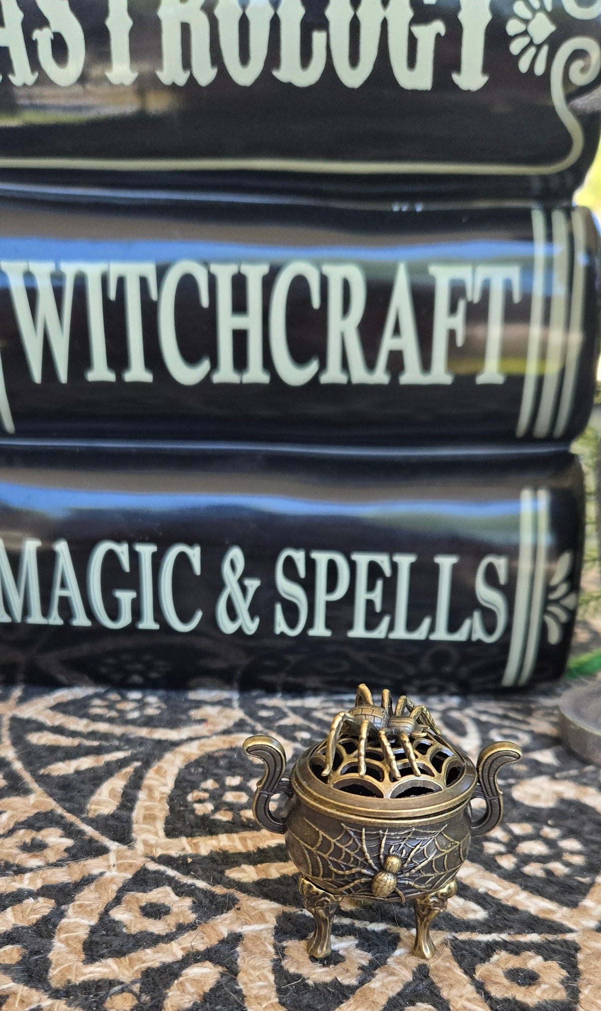 A small ornate bronze metal incense burner with a spiderweb design on the lid sits on a black and beige patterned cloth, with decorative books titled 'WITCHCRAFT', 'MAGIC & SPELLS', and 'ASTROLOGY' in the background.