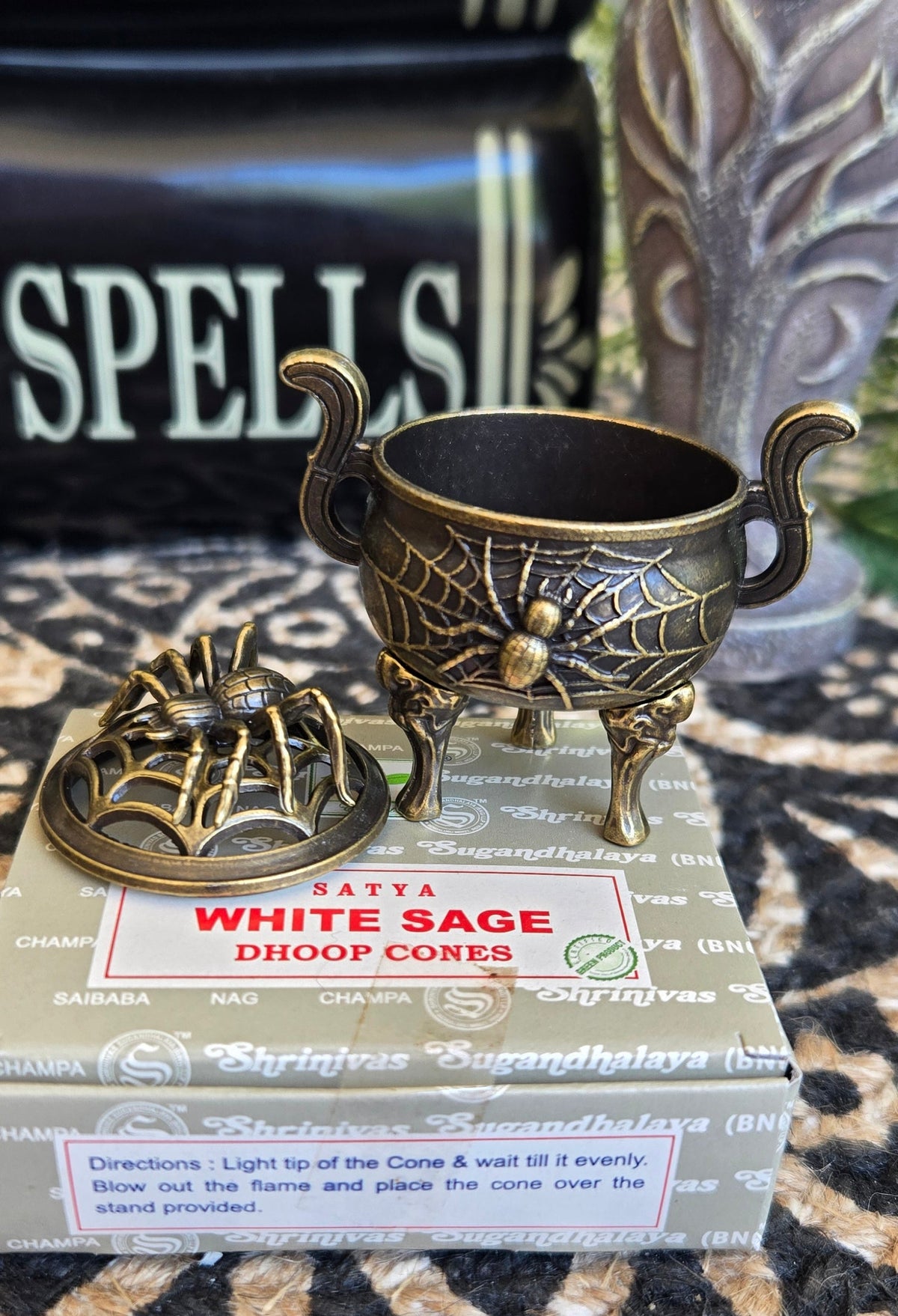 A brass cauldron incese burner on top of a white sage dhoop corner box, including a small tripod cauldron holder with spiderweb detailing, its matching lid, 