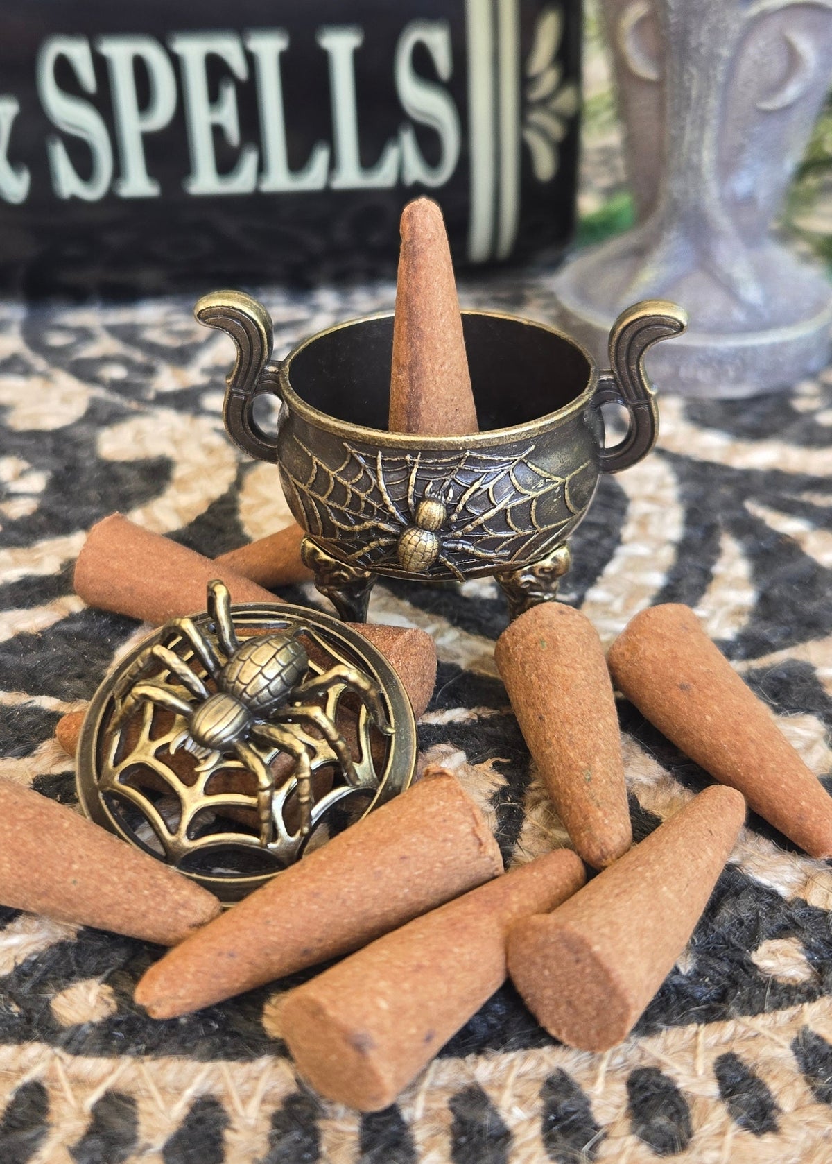 A small antique-style bronze cauldron adorned with a spider and its web beside the spider on top of a webbed lid. on a bed of cone incense