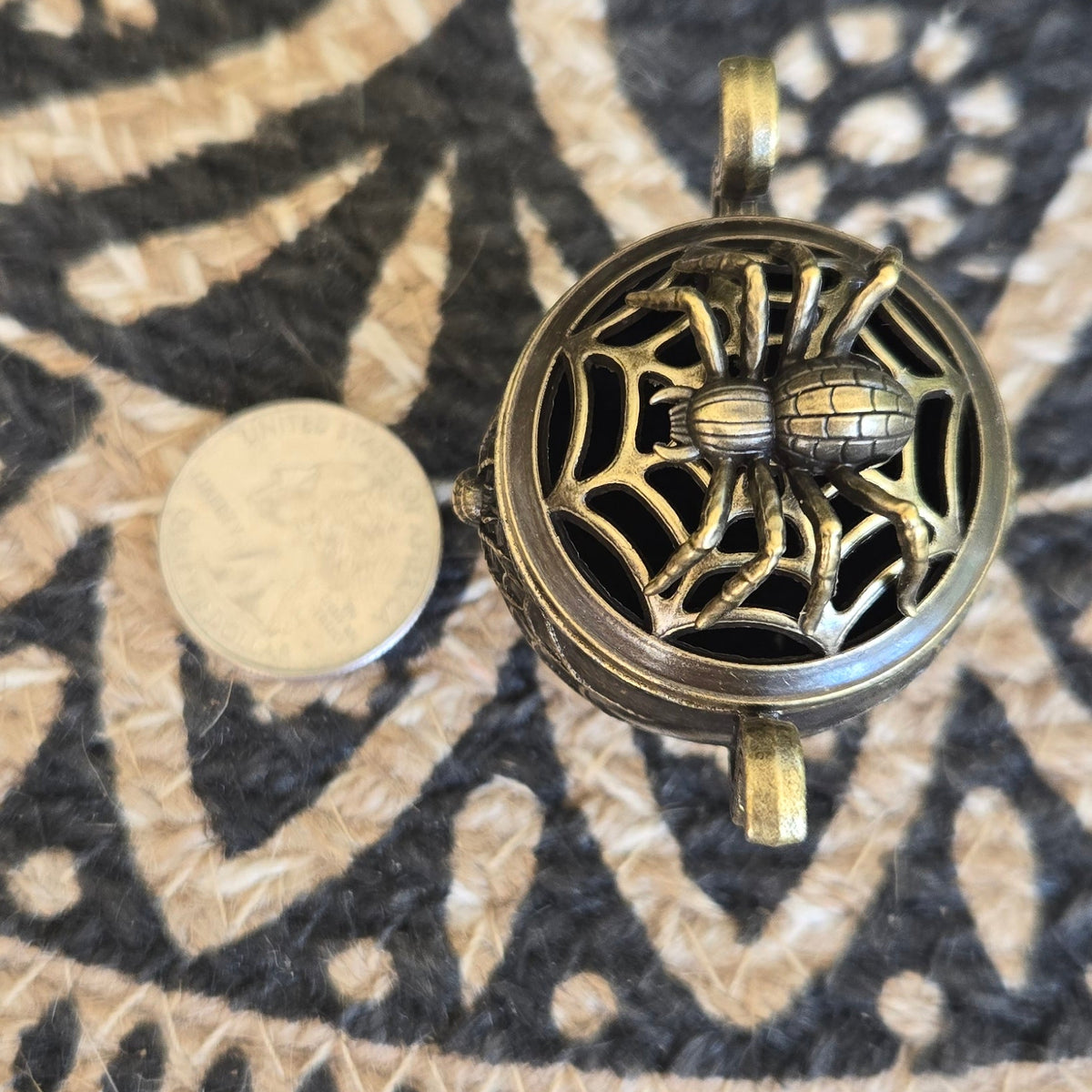 A round antique bronze metal pendant with an intricate filigree spiderweb design, shown next to a US Quarter coin for scale on a black and beige patterned rug.