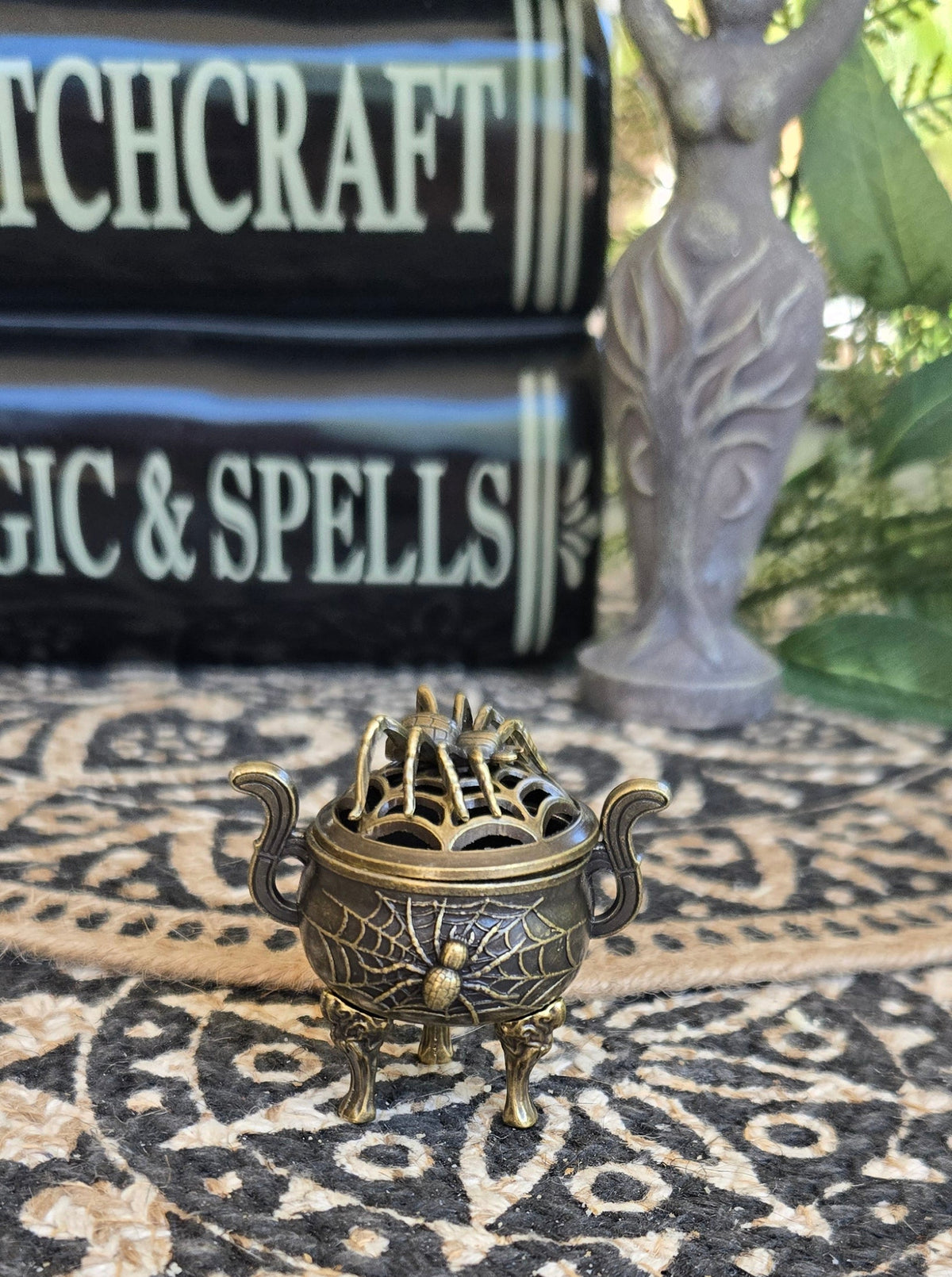A small ornate bronze cauldron incense burner with a decorative spider and web lid sits on a black and beige patterned rug, with books and a statue blurred in the background.