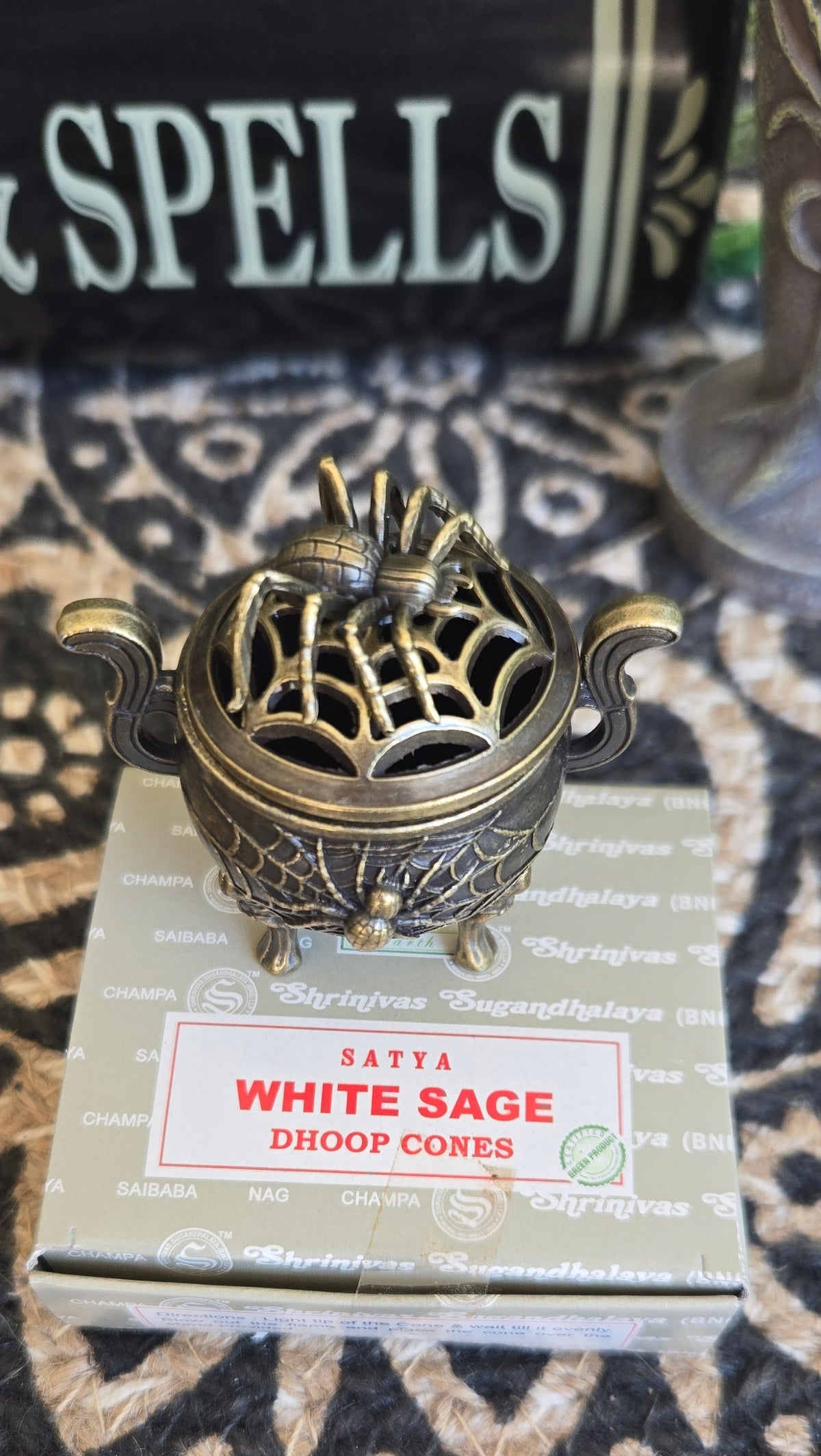 A small ornate metal cauldron adorned with spider and web incense burner sits on top of a box labeled 'Sattva White Sage Dhoop Cones' 
