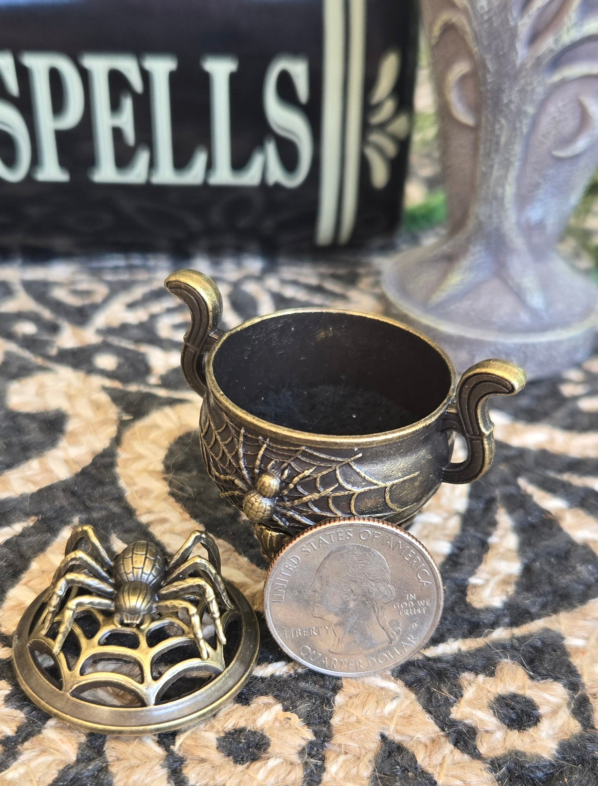 A small antique-style bronze spider-themed keepsake box with a lid featuring a spider cutout, shown next to a coin on a patterned rug with a book in the background.